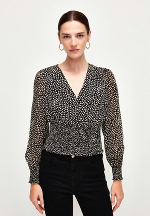 Black blouse with white polka dots, deep V-neck, long sheer sleeves, gathered waist, and smocked texture. Paired with black jeans.