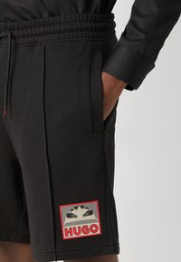 Shorts noirs avec une ceinture élastique, des poches latérales et un patch logo bordé de rouge comportant "HUGO" et un motif graphique. Texture de tissu doux.