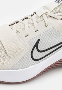 Scarpa sportiva beige con tomaia in mesh, caratterizzata da un logo Nike nero, un pannello testurizzato e una suola bianca con un accento rosso lungo il bordo.