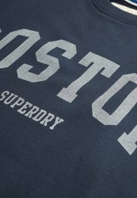 Sudadera azul marino con letras plateadas en relieve y de gran tamaño que dicen "BOSTON" y "SUPERDRY" en una fuente deportiva. Textura de tela suave.