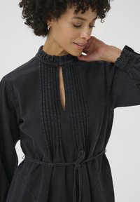Vestido de mezclilla negra con cuello con volantes, pliegues verticales y un corte en forma de llave, ceñido a la cintura con un cinturón de tela.