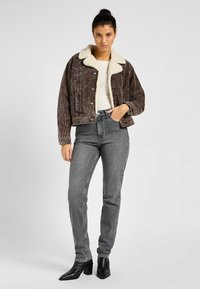 Chaqueta de pana marrón con un cuello de piel sintética color crema, cierre de botones y dos bolsillos, combinada con jeans grises de cintura alta y botas de tacón negras.