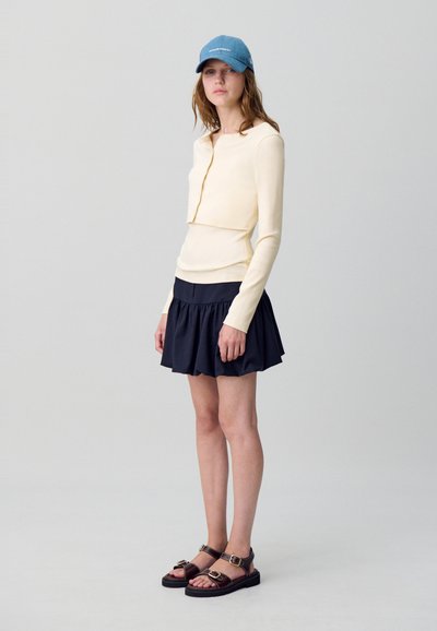 Jeune femme portant un haut crème à manches longues, une mini-jupe plissée marine, une casquette de baseball bleue et des sandales noires à boucles, se tenant devant un fond uni.