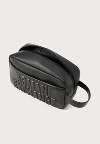 Črna usnjena torba s plišasto teksturo, z zadrgo inembosiranim logotipom "ARMANI EXCHANGE" spredaj. Ima zaobljeno obliko in ročaj.