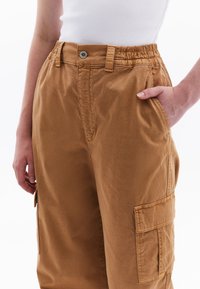 Cargo broek in lichtbruin katoenmix, voorzien van een elastische tailleband, voorvak en zijvak met klep. Gladde textuur.