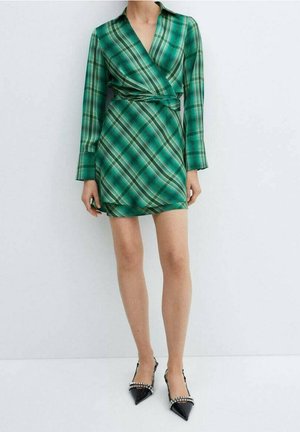 Kobieta ubrana w zieloną kratkę sukienkę z długimi rękawami i czarne szpilki z paskami ozdobionymi cyrkoniami, stoi na tle białego tła.