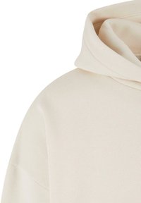 Sweat à capuche en tissu doux beige, montrant l'épaule et la capuche avec des coutures visibles et une texture, sur un fond blanc.