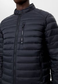 Svart vadderad pufferjacka med ståkrage, vertikal dragkedja på bröstfickan och två sidofickor med dragkedja, buren av en person med mörk hud.