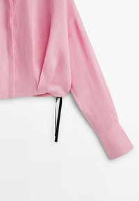 Chemise rose à manches longues avec larges poignets, dotée d'un cordon de serrage noir à l'ourlet inférieur sur fond blanc.