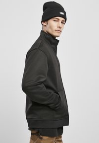 Juoda zip-up džemperis su aukštu apykakle, šoninėmis kišenėmis, raukšlėtais rankogaliais ir apačia. Dėvimas su tekstūruota juoda skrybėle ir kamufliažo kelnėmis.