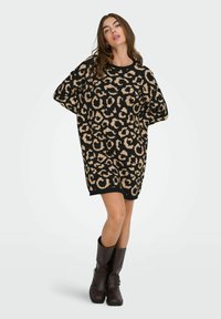 Robe tricotée avec un imprimé léopard en noir et beige, forme oversized, col rond et manches courtes ; associée à des bottes brunes jusqu'aux genoux.