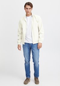 Lyse beige bomberjakke, lynlås lukning, elastiske manchetter, båret over en hvid t-shirt, parret med blå jeans og brune ruskindsko.