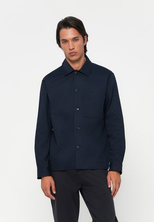 Young man wearing a navy button-up shirt with a chest pocket and dark trousers, standing against a plain white background.