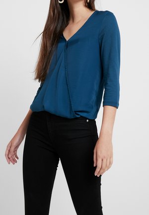 Femme portant un chemisier bleu à col en V avec des manches trois-quarts et un pantalon noir ajusté sur un fond uni.