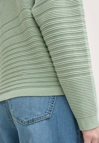 Suéter de punto acanalado verde con rayas texturizadas, que presenta un puño ajustado. Combinado con jeans de mezclilla azul claro, mostrando un detalle en el bolsillo.