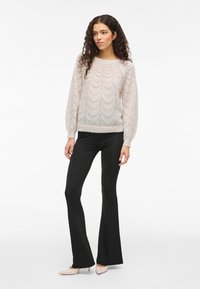 Γυναίκα με μακριά σγουρά μαλλιά, φορώντας ένα ελαφρύ πλεκτό πουλόβερ, μαύρα καμπάνα παντελόνια και μπεζ μυτερές γόβες, που στέκεται μπροστά σε λευκό φόντο.