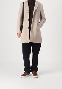 Beige wool coat with notched collar, two buttons, and a pocket. Worn over a brown ribbed sweater and paired with black pants and brown sneakers.