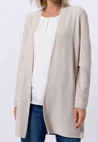 Cardigan beige texturé à devant ouvert et manches longues. Porté sur un chemisier blanc, associé à un jean bleu. Aucun bouton ni attache visible.