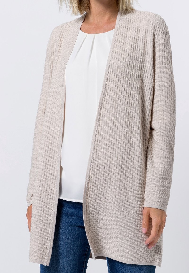 Cardigan beige texturé à devant ouvert et manches longues. Porté sur un chemisier blanc, associé à un jean bleu. Aucun bouton ni attache visible.