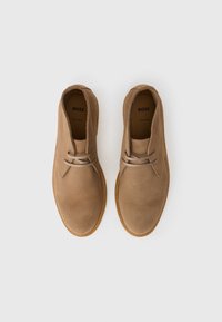Botine chukka din suede bej, cu vârful rotunjit, două găuri pentru șireturi și talpă cusută în contrast. Interiorul prezintă o branță cu marcaj.