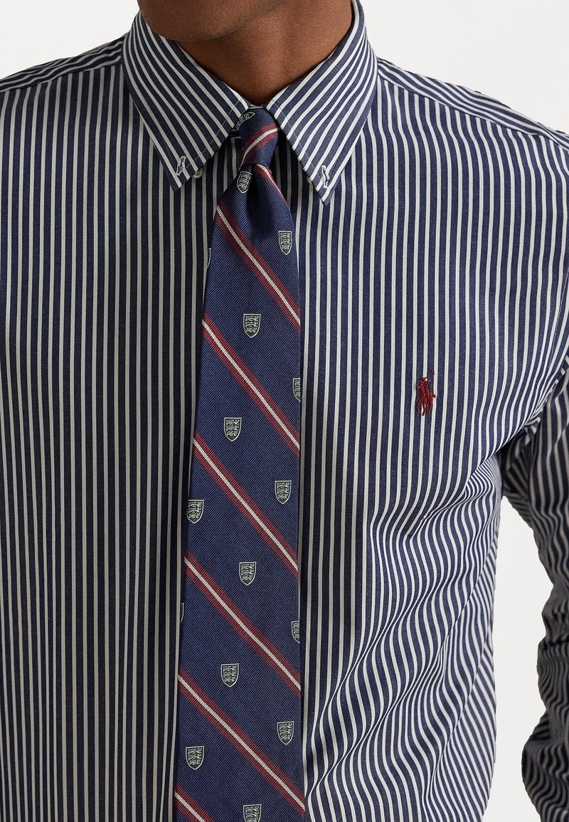Chemise à rayures bleu marine et blanche avec col à boutons, associée à une cravate bleu marine ornée de rayures diagonales rouges et de logos en forme de bouclier.