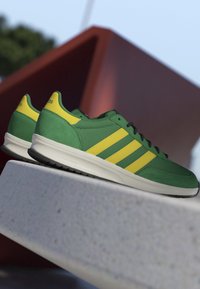 Green and yellow sneakers with black laces and white soles placed on a stone surface outdoors against a blurred geometric background.