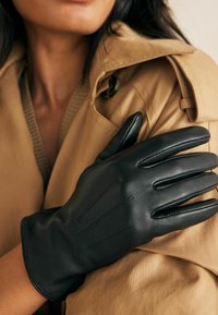 Guanti in pelle nera con finitura liscia, dotati di dita affusolate e cuciture dettagliate, indossati su una giacca beige con un colletto tessurizzato.