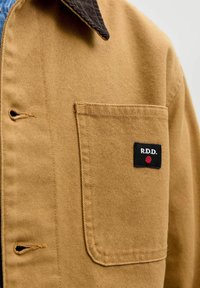 Close-up af tan jakkelomme med sort rektangulært mærke med teksten "R.D.D." og et rødt punkt under teksten, der viser syning og stoftekstur.