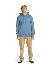 Sudadera de algodón azul con un bolsillo frontal, cordones y logo. Combinada con pantalones beiges y zapatillas en blanco y negro. Ajuste casual.