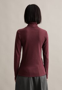 Maroon long-sleeve turtleneck top with a slim fit, ribbed texture, and a simple design, worn with a pleated gray skirt.