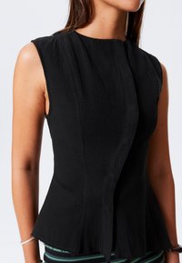 Top sans manches noir avec un tissu texturé, un corsage ajusté et un ourlet en peplum avec un léger chevauchement à l'avant.