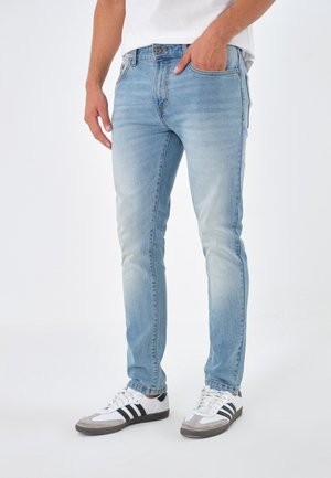 Homme portant un jean slim bleu clair, des baskets blanches à rayures noires et une chemise blanche, main dans la poche, debout sur un fond blanc.