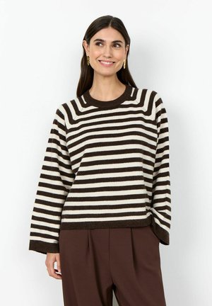 Nő barna és fehér csíkos, széles ujjú pulóvert és barna nadrágot visel, mosolyogva, kissé oldalra nézve.