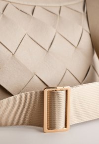 Detalj av en vävd läderväska i beige med en texturerad yta, bälte i guldfärgad metall och en bred tygrem.