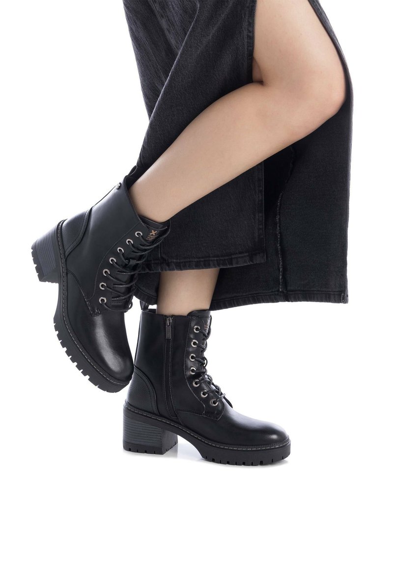 Botas de cuero negro hasta el tobillo con un tacón grueso, cordones frontal y cremallera lateral. Paneles texturizados y suelas de goma prominentes para mayor tracción.