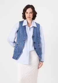 Colete de ganga azul com um design justo, botões frontais e bolsos laterais, usado sobre uma camisa às riscas azul e branca. Saia branca texturizada.