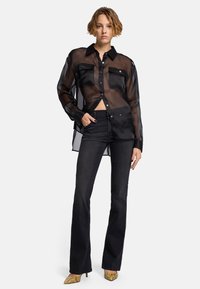 Schwarzes transparentes Hemd mit zwei Klappen-Taschen, kombiniert mit hochgeschnittenen schwarzen Schlaghosen und goldenen, spitz zulaufenden Schuhen.