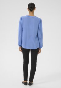 Svijetloplava bluza dugih rukava s nabranom teksturom i nabranim manžetama, u kombinaciji s crnim slim-fit hlačama i ravnim crnim cipelama.