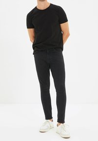 Man som står med händerna bakom ryggen, iklädd svart t-shirt, svarta slim fit-jeans och vita sneakers på vit bakgrund.