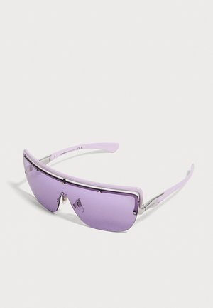 Lunettes de soleil futuristes enveloppantes avec des verres violets et des branches fines assorties sur un fond blanc uni.