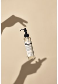 Transparent plastpumpflaska med svart pump och etikett. Innehåller mild rengöringsolja, svävande ovanför en skuggad hand, mot en beige bakgrund.