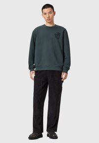 Tmavě zelená mikina s kulatým výstřihem a grafickým logem na hrudi, kombinovaná s černými manšestrovými kalhotami a černou obuví.