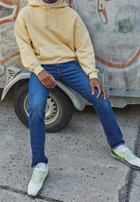 Sudadera beige con capucha y bolsillo frontal, jeans de mezclilla azules y zapatillas verde claro. Atuendo casual contra un fondo texturizado.