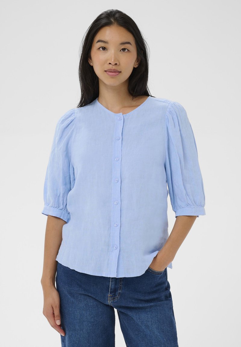 Femme aux longs cheveux noirs portant un chemisier bleu clair à boutons avec des manches bouffantes et un jean bleu, debout avec une main dans la poche.