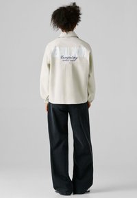 Persoon met krullend haar staat met de rug naar de camera en draagt een crèmekleurig jack met de tekst "Red Bull Racing Competing Since 2004" en zwarte wijde broek.