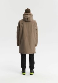 Osoba stojąca tyłem, ubrana w długi brązowy płaszcz z kapturem, czarne spodnie i szare buty turystyczne z jasnoniebieskimi podeszwami.