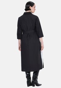 Abito camicia lungo nero con colletto, vita cinturata, maniche a ¾ e spacchi laterali. Tessuto liscio e leggero con un colore uniforme.