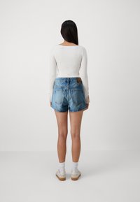 Shorts di jeans di colore blu chiaro con orlo sfilacciato, cinque tasche e un'etichetta in pelle marrone. Parte di una maglietta bianca a maniche lunghe è visibile.