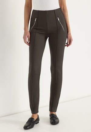 Slim-fit mørkebrune bukser med front lodrette syninger og lynlåslommer, båret med sorte loafers og en hvid ærmeløs top.