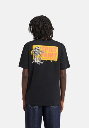 Personne avec des dreadlocks porte un t-shirt noir montrant un squelette de dessin animé tenant un pinceau assis sur une boîte de peinture avec le texte "Spilt Paint."